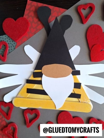 A bee-themed gnome made from colored paper is surrounded by red felt hearts on a craft background, perfect for a Valentine's Day bee or popsicle stick bee gnome project. A label at the bottom right reads @GLUEDTOMYCRAFTS.