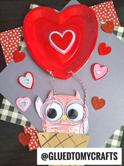 A red paper plate hot air balloon floats above a paper owl with big googly eyes in a basket. Red and pink heart decorations are scattered on a patterned background. Text reads @GLUEDTOMYCRAFTS.