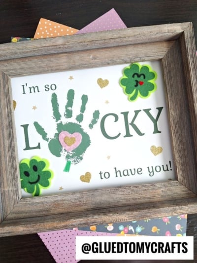 Framed artwork with the phrase I'm so LUCKY to have you! featuring a green handprint lucky U, surrounded by felt shamrocks, hearts, and gold glitter accents on colorful polka dot paper.