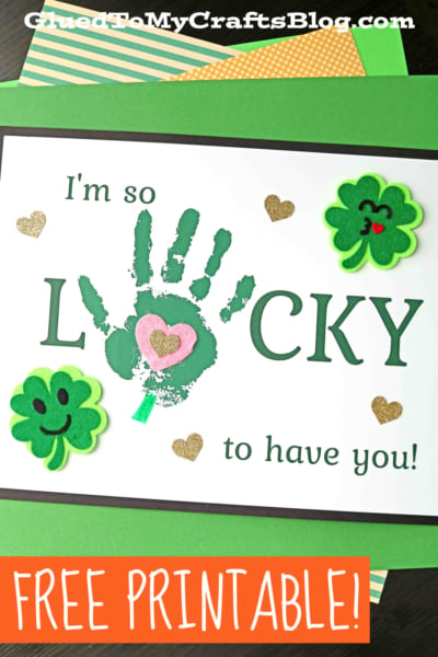A St. Patrick’s Day card says “I’m so lucky to have you!” with a green handprint forming the "U," surrounded by shamrock stickers and glittery gold hearts—perfect for a handprint lucky keepsake. The bottom reads “FREE PRINTABLE!”.