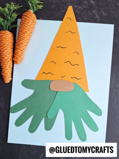 A carrot gnome paper craft made from cut-out shapes: an orange triangle hat, two green hand shapes as a beard, and a brown oval nose on a light blue background. Two carrot decorations are placed beside the craft.
