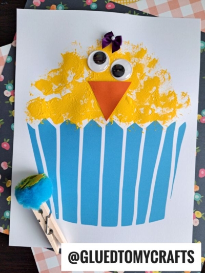 A cute easter chick cupcake craft made with a blue-striped liner, yellow paint, googly eyes, an orange beak, and a purple ribbon on top. A blue pompom clothespin sits beside it. @GLUEDTOMYCRAFTS label at the bottom.