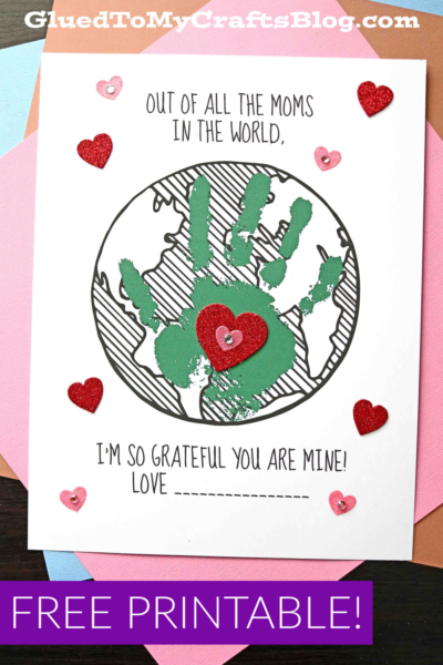 A handmade Mother’s Day card features a green handprint on a globe, surrounded by red and pink glitter hearts. This mom world keepsake reads: “Out of all the moms in the world, I’m so grateful you are mine! Love __.”.