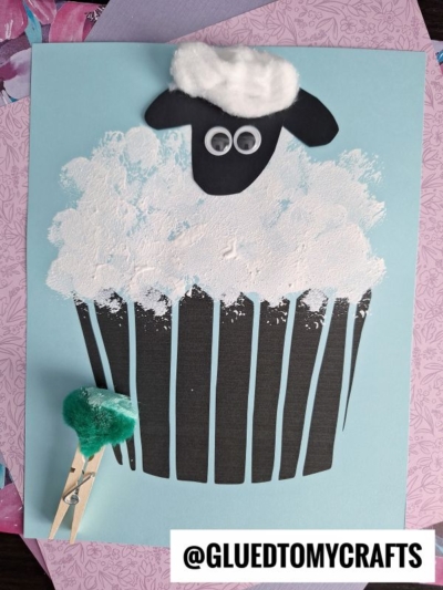 A sheep cupcake craft featuring black paper, white cotton ball frosting, and googly eyes sits on a blue background. A green pom-pom attached to a clothespin accents the lower left. Text reads @GLUEDTOMYCRAFTS.