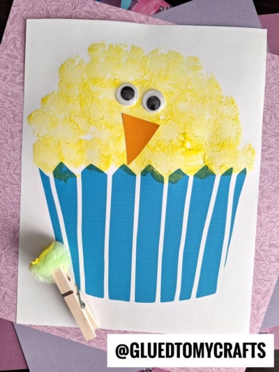 A childrens craft resembling an Easter chick cupcake, with a yellow chick face, blue wrapper, googly eyes, and orange beak—made using paint and a pom-pom on a clothespin. @GLUEDTOMYCRAFTS watermark at the bottom.