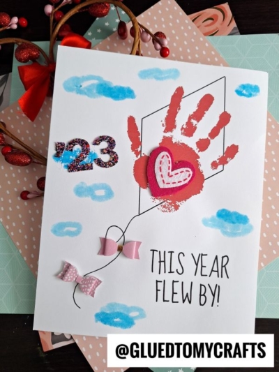 A handmade card features a red handprint forming a kite, embellished with a pink heart, pink bows for the tail, glittery “23,” blue clouds, and the phrase “This year flew by!” @GLUEDTOMYCRAFTS is at the bottom.
