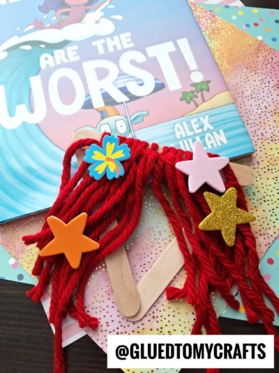 A handmade craft featuring vibrant yarn mermaid hair and a red-haired tail, created with foam star stickers and craft sticks, sits atop colorful paper and a children’s book titled ARE THE WORST! by Alex Willan.