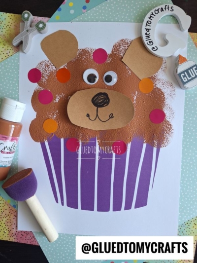 A children’s bear cupcake craft features a cupcake with brown paint, googly eyes, and colored paper on a patterned background. Craft supplies like glue, paint, sponge, and scissors surround the adorable bear cupcake masterpiece.