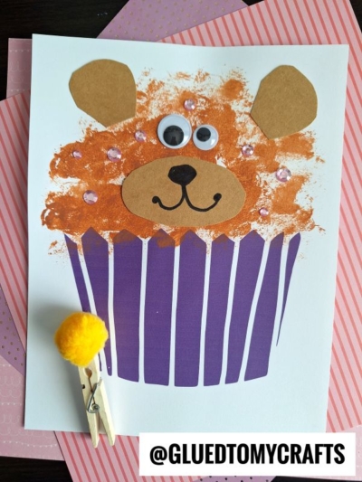 A bear cupcake craft made with a purple paper cupcake liner, brown paper ears and snout, googly eyes, pink gems, and orange paint. A clothespin with a yellow pom-pom is beside this adorable bear cupcake creation.