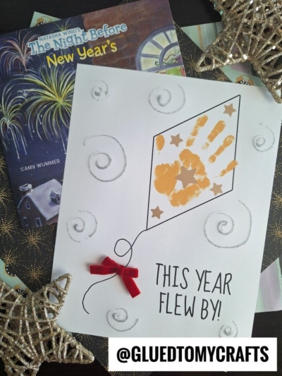 A children’s craft features a handprint as the center of a paper kite with "The year flew by!" written below. A small red bow decorates the kite string, swirls add flair, and The Night Before New Years book sits nearby.