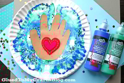A paper plate painted with blue and green swirls creates a paint splat earth effect. A brown paper hand with a red heart is glued in the center, surrounded by paint bottles, confetti, and craft paper.