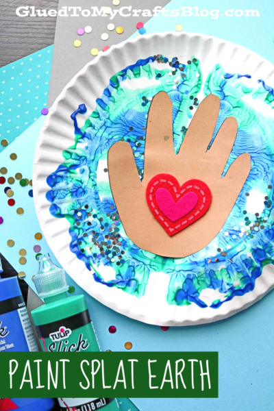 A paper plate painted with blue and green swirls creates a paint splat earth effect. A brown paper hand with a red heart is glued in the center, surrounded by paint bottles, confetti, and craft paper. Text reads Paint Splat Earth.
