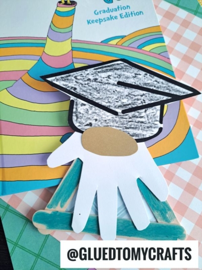 A homemade graduation craft featuring a popsicle stick graduation gnome with a paper handprint and a colored cap sits atop a vibrant Graduation Keepsake Edition book. @GLUEDTOMYCRAFTS is written at the bottom.