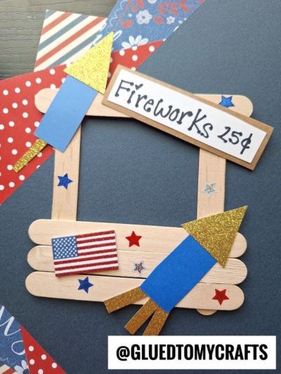 A popsicle stick firework stand photo frame, decorated with paper rockets, glitter, an American flag sticker, stars, and a “Fireworks 25¢” sign. Red, white, and blue patterned paper completes this festive DIY craft.