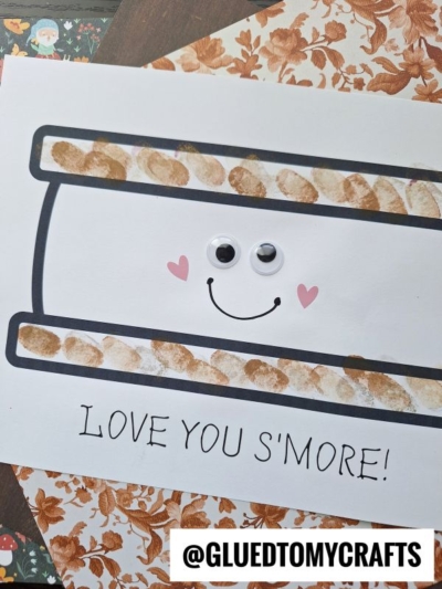 A cute fingerprint s'mores craft made with brown paint thumbprints for graham crackers, two googly eyes, a smiling face, and pink heart cheeks. Text below reads LOVE YOU SMORE! and @GLUEDTOMYCRAFTS is at the bottom.