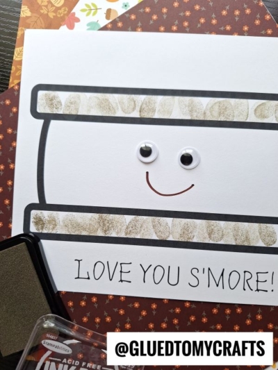 A handmade s’more card featuring fingerprint s’mores with googly eyes, a smile, and LOVE YOU SMORE! written at the bottom. Decorative paper and a black ink pad are nearby.