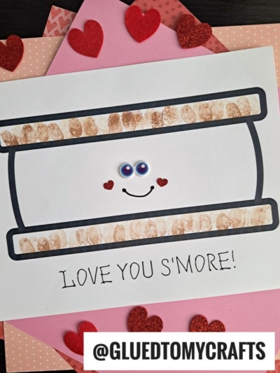 A handmade Valentines card with a drawing of fingerprint s'mores, googly eyes, a smiling face, and heart cheeks. The caption reads “LOVE YOU S’MORE!” Red glitter hearts and pink paper decorate the background.