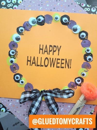 An orange card with Happy Halloween! in the center, decorated with a Halloween eyeball wreath made of painted circles, googly eyes, a black and white bow, and a small orange pompom craft tool in the corner.