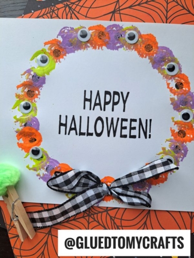 A Halloween eyeball wreath with colorful painted dots, googly eyes, and a black-and-white gingham bow surrounds the words HAPPY HALLOWEEN! on white paper. A green pom-pom and clothespin add a playful touch to this festive craft.