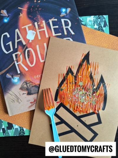 A children’s book titled Gather Round sits beside a fork painted campfire craft, featuring vibrant flames created on brown paper. The handle @GLUEDTOMYCRAFTS appears at the bottom.