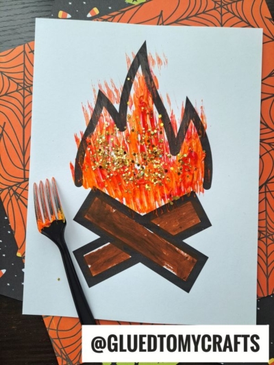 A fork painted campfire craft with vibrant, glitter-accented flames and crossed paper logs. An orange-painted fork rests beside it. GLUEDTOMYCRAFTS is printed at the bottom.