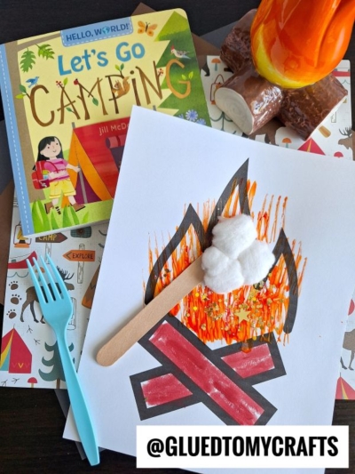 A children's camping-themed craft scene with a book titled Let's Go Camping, a drawing of a campfire decorated with orange lines, cotton balls, a craft stick, plastic utensils, and a fork painted campfire on a patterned surface.