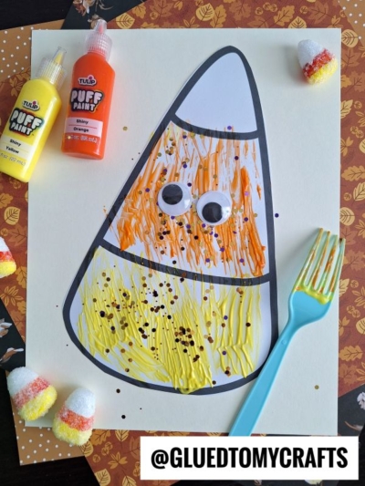 A fork painted candy corn craft with googly eyes features orange and yellow puffy paint. A blue plastic fork, paint bottles, and candy corn candies rest on a fall-themed background.