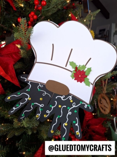 A handmade Christmas ornament shaped like a chef's hat with holly, attached to black paper handprints decorated with sequins, creates a festive handprint chef gnome hanging on the tree. The handle @GLUEDTOMYCRAFTS is at the bottom.