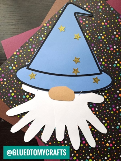 A handprint wizard gnome with a blue hat decorated with gold stars and a white beard made from handprints, placed on star-patterned paper.