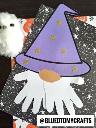 A handprint wizard gnome craft with a purple wizard hat, gold stars, and a white handprint beard sits on star-patterned paper. A small white owl plush is beside it.