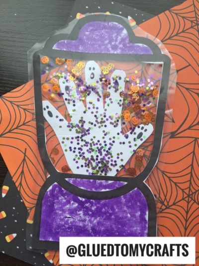 A Halloween-themed craft featuring a handprint inside a blender outline, filled with sparkly pumpkin and circle confetti, resembling a stained glass Halloween lantern, displayed on spiderweb and candy corn patterned papers.