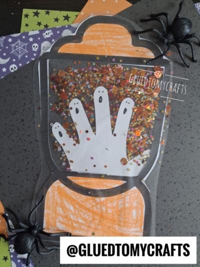 A Halloween craft features a paper jar with a white handprint, orange confetti, and spooky decorations like fake spiders and patterned paper, creating the look of a stained glass Halloween lantern. The handle @GLUEDTOMYCRAFTS is displayed.