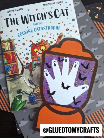 A children’s book titled The Witch’s Cat and the Cooking Catastrophe lies partly under a Halloween-themed craft shaped like a crystal ball with a white handprint, black bats, and a stained glass Halloween lantern on a purple background.