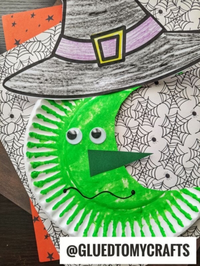 A green paper plate witch with a googly eye, paper nose, and large hat sits on spider web patterned paper beneath a shimmering moon. @GLUEDTOMYCRAFTS is written at the bottom.