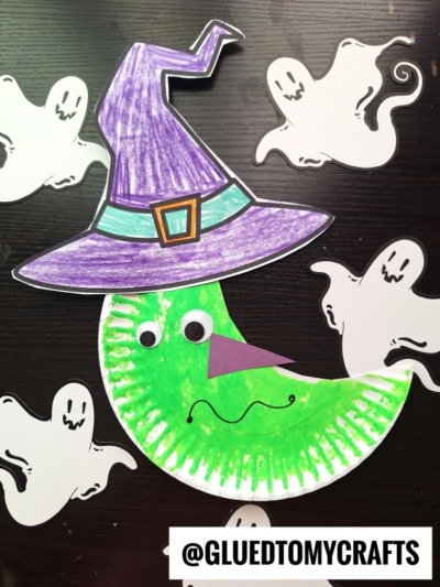 A paper plate witch moon craft made with a green crescent moon wearing a purple witch hat, googly eyes, and surrounded by five white paper ghosts. The text @GLUEDTOMYCRAFTS appears at the bottom.