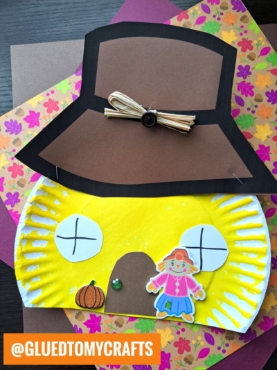 A colorful craft featuring a yellow paper plate with a pilgrim hat, a pumpkin, a girl, and fall-themed paper in the background creates a cozy paper plate scarecrow house scene. The handle @GLUEDTOMYCRAFTS appears on the bottom corner.