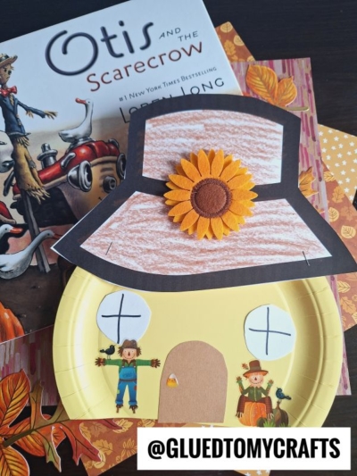 A children’s craft featuring a paper plate scarecrow house with a large brown hat and sunflower, placed on autumn-themed paper next to the book Otis and the Scarecrow. The handle @GLUEDTOMYCRAFTS is displayed.