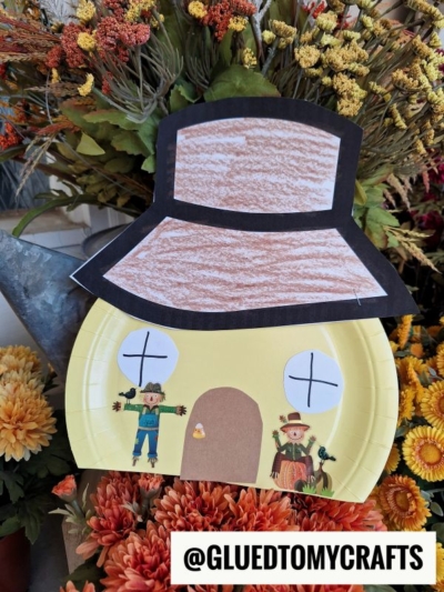 A paper plate scarecrow house decorated as a yellow home with drawn-on windows, door, and roof, plus two scarecrow stickers. The house is surrounded by orange and yellow flowers. @GLUEDTOMYCRAFTS is written at the bottom.