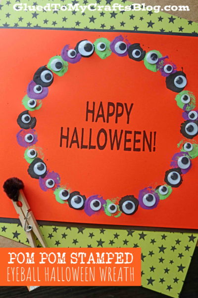 An orange card with HAPPY HALLOWEEN! in black, surrounded by a Halloween eyeball wreath made from colorful pom-pom stamped eyeballs. The card sits on green star-patterned paper, accented with a clothespin and pom-pom in the corner.