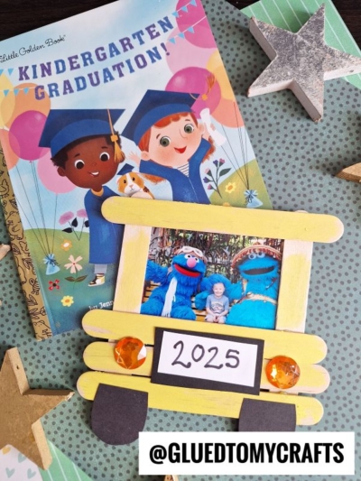 A “Kindergarten Graduation” book with illustrated children in caps lies beneath a yellow school bus frame craft holding a photo and “2025” sign, surrounded by glittery gold and silver stars on a polka dot background.