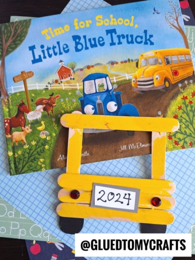 Childrens book Time for School, Little Blue Truck with a yellow school bus craft made from popsicle sticks, red gems, and a license plate labeled 2024 on patterned paper. The school bus frame adds a playful touch to this creative project. @GLUEDTOMYCRAFTS watermark at bottom.