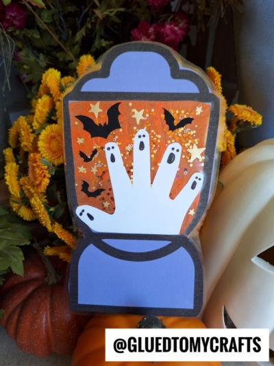 A Halloween craft features a paper handprint decorated as ghosts, surrounded by bats and stars, set against an orange background. Yellow flowers, pumpkins, and a stained glass Halloween lantern complete the festive scene. Text reads @GLUEDTOMYCRAFTS.