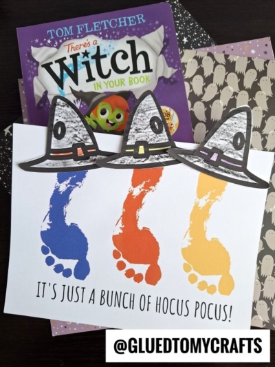 A children’s book titled There’s a Witch in Your Book sits with Halloween papers, including one with ghost patterns. In front, a hocus pocus keepsake craft shows footprint Sanderson sisters’ hats above painted footprints and the words, “It’s just a bunch of hocus pocus!”.