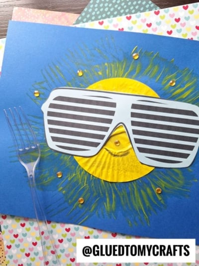 A craft project of a smiling fork painted sun made from a yellow cupcake liner on blue paper, wearing large striped sunglasses, with yellow paint rays and decorative gems. A plastic fork and patterned heart paper are nearby.