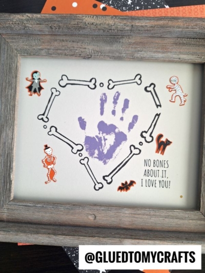 A framed Halloween keepsake featuring a purple handprint inside a heart of bone illustrations, surrounded by Halloween stickers, with the text “No bones about it, I love you!”—perfect for spooky season memories.