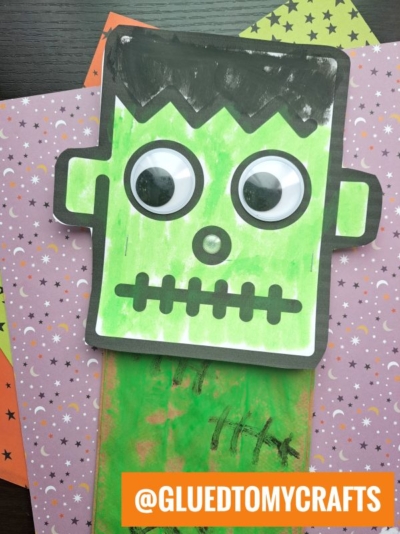 A green paper bag Frankenstein puppet with big googly eyes, black hair, and a stitched mouth is set on colorful star and moon patterned paper. An orange box reads @GLUEDTOMYCRAFTS at the bottom.