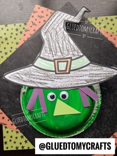 A handmade paper bowl witch craft features a green-painted plate, a paper hat, googly eyes, and colorful paper shapes for the nose and hair, all displayed on patterned paper.