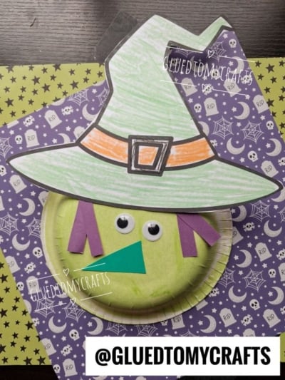 A paper bowl witch craft with a green hat, googly eyes, and purple and green paper features sits on patterned Halloween paper with skulls, spiders, and webs. @GLUEDTOMYCRAFTS is written at the bottom.