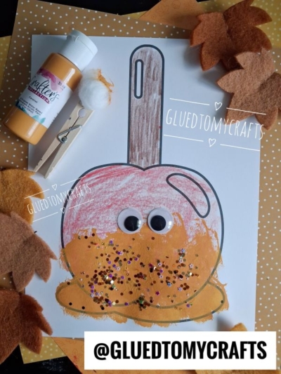 A stamped caramel apple paper craft with googly eyes, decorated with glitter, felt leaves, paint, and a pom-pom, sits on a polka-dot background. The text @GLUEDTOMYCRAFTS is shown at the bottom.