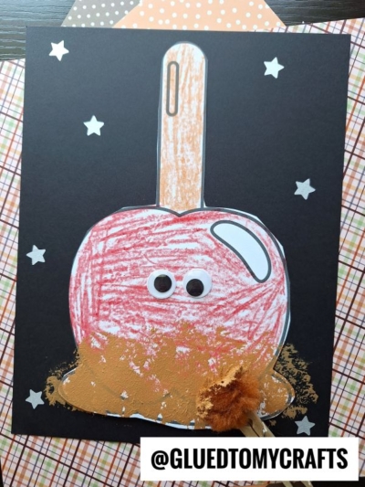 A kids craft of a stamped caramel apple on black paper with stars, googly eyes, and a real popsicle stick. Crayon coloring and brown paint mimic caramel. Background has plaid and polka dot paper. @GLUEDTOMYCRAFTS text at bottom.